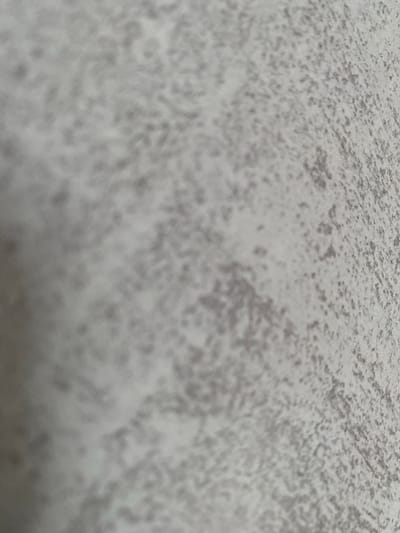 The image is a close-up, shallow-depth-of-field photograph of a textured, light gray surface. The texture appears granular or speckled, with darker gray or purplish-gray speckles distributed unevenly across the surface. The lighting creates subtle shadows and highlights, suggesting the surface has a three-dimensional quality. The photograph is taken at an angle, with the left side of the image appearing more out of focus and the right side slightly sharper. There are no people, objects, or discernible environmental features visible that would allow for the identification of a specific location, including Александровка, Russia.