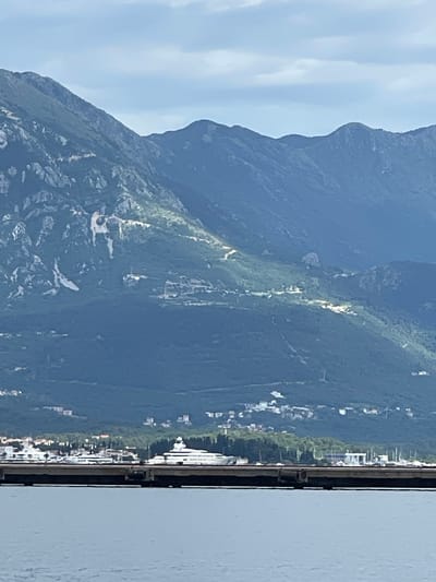This horizontal landscape photograph captures a scenic view of the mountainous coastline of Bijela, Montenegro. The scene is dominated by a majestic, dark green mountain range that rises steeply from the water's edge, stretching across the middle and left portions of the frame. The mountain slopes are covered in dense vegetation, with scattered lighter patches and streaks, possibly indicating exposed rock formations, sparse growth, or trails carved into the terrain.
Above the mountain, the sky is a clear, light blue with soft, wispy white clouds, suggesting a pleasant daytime setting. In the foreground, on the right side of the image, a calm body of water is visible, likely the Bay of Kotor, extending from the bottom to the top of the frame. A dark, vertical structure, possibly a railing from a boat or a waterfront promenade, runs along the immediate right edge, framing the view.
Along the base of the mountains, nestled between the water and the rising slopes, a coastal settlement is visible. Numerous white and light-colored buildings are scattered amidst green areas, indicating a vibrant town or village. No specific activity or individuals are discernible, presenting a tranquil and expansive landscape. There is no visible text in the image.