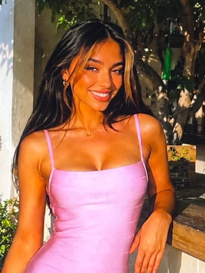 A young woman with long, dark, layered hair with blonde highlights is smiling and leaning on a wooden surface outdoors. She is wearing a strapless pink dress with thin straps and a delicate gold necklace. Her skin is tanned, and she has gold hoop earrings. The background is blurred, showing foliage and a light-colored wall. The lighting suggests it is daytime, possibly late afternoon due to the warm tones. The setting appears to be a cafe or restaurant with an outdoor seating area, consistent with many establishments in the Plaka district of Athens, Greece, known for its charming ambiance and picturesque surroundings.