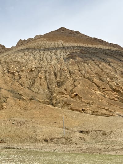 A landscape photograph, oriented vertically, shows a large, multi-layered, heavily eroded rock formation beside a body of water. The rock formation features horizontal striations and deep vertical fissures. The ground near the water's edge is dry and sparsely vegetated. There is a thin, metallic object, possibly a pipe or rod, running along the ground parallel to the water. The sky is visible in the upper left corner and is a pale, overcast blue. The image is located in Tashi Dzom town, China.