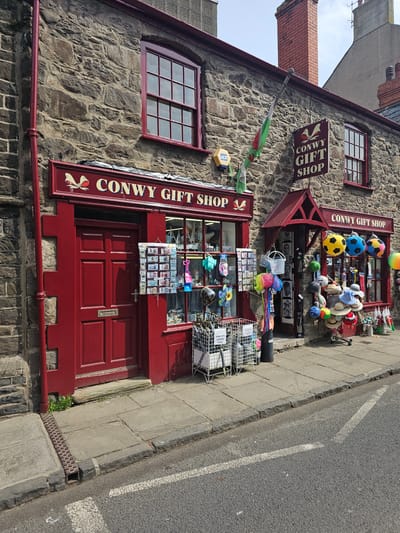 The image depicts the facade of the "Conwy Gift Shop," a two-story building with stone walls and red trim, located in Llandudno Junction, United Kingdom. The shop's signage is prominently displayed on both floors. Souvenirs, including balls, hats, and postcards, are exhibited in front of the shop windows and entrance. An ADT security sign is visible on the wall. A Welsh flag hangs near the upper window. The adjacent pavement and road are visible, indicating an urban setting.