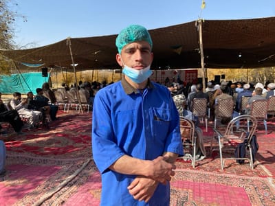 A young adult male stands in the foreground, facing the camera, within an outdoor gathering in Sard-E 'olya, Afghanistan. He wears a blue short-sleeved uniform shirt, a light blue disposable hairnet, and a light blue surgical mask pulled down below his chin; his hands are clasped in front. Behind him, under a large brown tarp canopy supported by wooden poles, numerous adult males are seated on metal chairs arranged in rows on red Persian-style rugs. Attendees are dressed in traditional and casual attire. A white banner with red markings is visible in the distant background under the canopy, along with speaker equipment to the left. The scene is illuminated by natural daylight under a clear blue sky, with trees visible beyond the canopy in the far background.