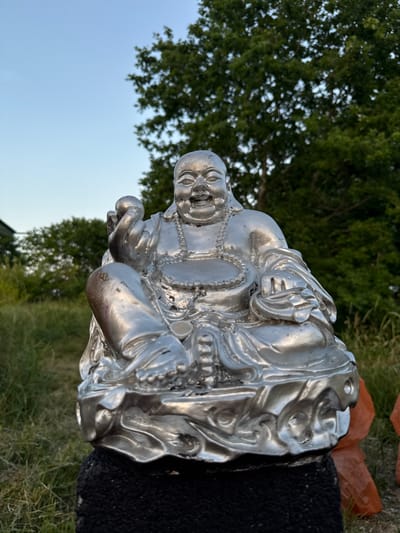 A silver statue of a laughing Buddha sits outdoors in Vol'noe, Russia. The statue is on a dark, textured pedestal. The Buddha is depicted in a seated position with one hand holding a round object and the other resting on its lap. It wears a beaded necklace and ornate robes. The background consists of green grass, trees, and a clear blue sky.