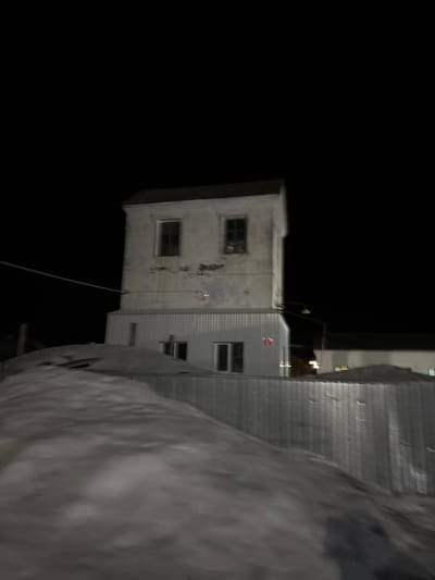 The image presents a nocturnal scene in Bayronovka, Russia, dominated by a two-story building under a dark, clear sky. The foreground is occupied by a large, undulating mound of snow, obscuring the ground level. Behind the snow, a corrugated metal barrier or fence spans horizontally.
The central building features an older upper story with weathered, light-colored walls and a dark, sloped roof. Two rectangular windows with dark panes are visible on this upper section. Below, the newer lower story is clad in vertical corrugated metal siding, appearing silver or light grey. This lower section extends horizontally beyond the upper structure and includes at least two windows: one narrow vertical and one wider horizontal. A small, faint red object or light source is present on the lower facade, slightly right of center. A thin, dark cable extends diagonally from the upper left of the frame toward the upper story of the building.
To the right of the main building, a street lamp on a pole provides limited illumination to the background, revealing the dark outlines of additional structures. No individuals, animals, or vehicles are present. No discernible actions or interactions are depicted.