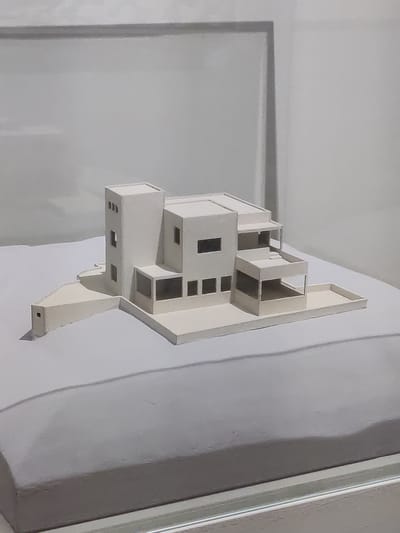 This is a close-up, medium shot of an architectural model of a multi-story building. The model is made of white material and is mounted on a larger white surface. The building has a geometric design with stacked rectangular volumes, some of which appear to have open sections or balconies. There are various rectangular openings on the model, representing windows or doorways. The lighting in the image is soft and diffused, suggesting an indoor setting with artificial light. The background is a plain, light-colored wall or surface, with some subtle shadows and lighter areas that create a sense of depth. The perspective is from a slight angle, allowing a view of the three-dimensional form of the model. No people, text, or specific time of day or weather cues are visible. The context provided indicates the location is The Hague, Netherlands, which is a cue to the potential style or origin of the architectural design, but this cannot be confirmed from the image alone.