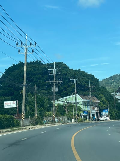 The image depicts a street scene in Baan Chaweng Noi, Thailand, under a clear blue sky. A paved road curves through the frame, marked with white dashed lines and a prominent yellow center line. On the left side of the road, a series of power poles with numerous wires are visible, supporting electrical infrastructure. A sign for "ROSE GARDEN BANGALOW" is mounted on a pole. Behind the road and power poles, a densely vegetated hill covered in lush green trees forms a prominent backdrop. Several buildings are situated along the roadside, including a pale green structure with multiple balconies and windows, and a white car parked in front of one of the buildings. The environment appears to be a semi-urban or suburban area with a mix of infrastructure, natural elements, and commercial establishments.