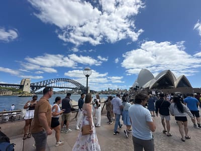 The Sydney Opera House and Harbour Bridge dominate a scene filled with tourists. A clear blue sky with scattered clouds provides a bright backdrop. Diverse individuals dressed in casual summer attire stroll along a promenade. The iconic architecture and the bridge suggest a popular sightseeing location, bustling with activity on a sunny day.