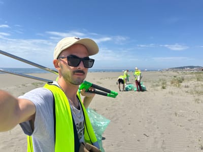 The image shows a man wearing a neon yellow safety vest, a grey shirt, a light-colored cap, and dark sunglasses. He is holding a green and black reaching tool, presumably used for picking up litter. In the background, there are other people also wearing safety vests who appear to be collecting trash. They are on a sandy beach, and there is the sea and horizon line in the distance. The photo seems to be taken as a selfie. The location appears to be the beach near Donji Štoj, Montenegro, based on the geographical features of the coastline and the nature of the beach environment.