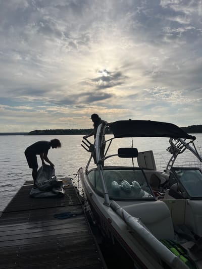 A waterside scene captures two individuals near a boat docked at a pier. One person stands on the dock, holding a large, bundled object wrapped in a reflective material. The other person is on the boat, possibly adjusting equipment or the boat's tower. The boat is moored to the wooden pier, which extends into the water. The sky is filled with clouds, with the sun shining through in the background. In the distance across the water, there is a treeline. Given the landscape and context clues, it is most likely that the image was taken in a rural area in a place such as Пятница, Russia, but there are no identifiable details from the city present in the image.