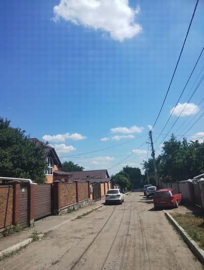 This image depicts a street scene in Sumy, Ukraine, on a clear day with scattered white clouds. The perspective is angled upwards, capturing a bright blue sky with overhead power lines and a utility pole.
The foreground shows a residential area with brick fences and houses. One house has a distinctive brown tiled roof and a white-framed window. Adjacent to the houses are two large, brown garage doors. Trees with dense green foliage line the properties.
The street itself appears to be a dirt or gravel road, with a concrete curb running along one side. Several cars are parked or driving on the street. A red hatchback is parked on the side of the road, and further down, a white car is visible, followed by another lighter-colored vehicle. The cars have visible license plates, although the text is not clearly legible.
The time of day appears to be daytime, likely late morning or early afternoon, given the bright sunlight and shadows. The weather is pleasant and sunny, conducive to outdoor activities. The scene conveys a sense of everyday life in a suburban or semi-rural part of Sumy. There is no text that is clearly legible in the image.