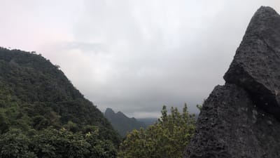 The image depicts a lush, mountainous landscape under an overcast sky, likely Vang Vieng, Laos, a region known for its karst topography. In the foreground, on the right side, a large, jagged rock formation dominates the frame, its rough texture highlighted. To the left, a densely forested mountain slopes upwards, covered in a thick canopy of green trees. In the middle ground, a valley is visible with more jagged peaks piercing through a layer of mist. The vegetation is verdant and appears to be a tropical rainforest. The overall mood is subdued due to the cloudy weather, creating a sense of depth and scale in the natural environment.
