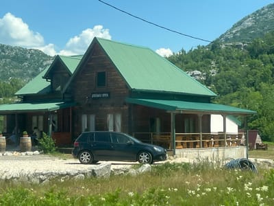 This is a medium shot, eye-level, outdoor photograph of a dark blue Volkswagen Golf hatchback parked on a gravel area in front of a wooden, two-story building with a green corrugated metal roof. The building appears to be a rustic lodge or restaurant, with a covered porch area featuring wooden tables and benches. Text in Cyrillic script is visible above a window on the building's facade. To the left of the building, two wooden barrels are present. In the background, lush green mountains covered in trees rise under a bright blue sky with scattered white clouds. The foreground consists of dry grass and wildflowers. The scene is brightly lit, suggesting it was taken on a sunny day. No specific actions or interactions are clearly depicted, but two individuals are faintly visible sitting on the porch on the left side of the building. The image appears to be taken in Dragalj, Montenegro, given the context provided.