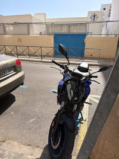 The image shows a blue and black motorcycle parked on an asphalt surface in what appears to be an outdoor area, possibly a street or a parking lot, in Ibiza, Spain. The motorcycle is the primary subject, positioned in the foreground and angled towards the viewer. It's a daytime shot, with bright sunlight casting shadows, suggesting clear weather.

In the background, a building with beige and white facades is visible. A blue metal door or panel is part of the building's architecture, with a metal fence running horizontally in front of it. Above the building, a clear blue sky is visible. A sign with text and a circular emblem is mounted on the building's white facade. The emblem features a graphic of hands holding something and text around it, which is partially obscured.

To the lower right of the motorcycle, the rear of a light-colored car is visible, featuring a license plate with the text "091 DVD". The car's taillight and Audi logo are also discernible. The ground is asphalt with some yellow markings. A metal beam or railing runs along the top right of the image, creating a frame. The overall scene is static, with no people or discernible activity taking place, presenting a typical urban or semi-urban environment in daylight.