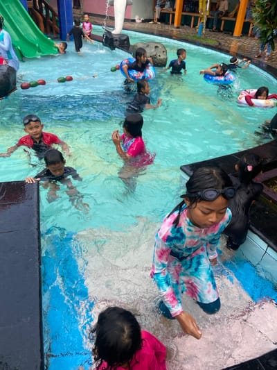 The image shows a swimming pool with multiple children swimming and playing in the water. The pool is located in the city of Kecamatan Tanggulangin, Indonesia, with coordinates of latitude -7.497 and longitude 112.684. The pool is surrounded by a concrete deck and a small area with grass and plants. There is a slide and a small waterfall entering the pool. There is also a rope with floating balls in the pool, and some children are using inflatable tubes. The children are wearing various swimming attire, including swimsuits, rash guards, and floaties. Some of them are interacting with each other while playing in the water.  There is a girl in a pink and blue patterned swimsuit standing on the edge of the pool with her hand outstretched towards the water. Behind her, a group of children are enjoying the swimming pool.  Some adults are sitting on the edge of the pool watching the children, but they are not visible in the picture. The pool is surrounded by a concrete wall and a fence. There is a small, light-colored building and a small garden nearby. The pool itself has a blue, tiled surface and is filled with clear water.