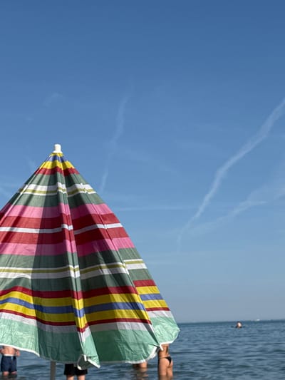 The image depicts a beach scene with a focus on a striped beach umbrella. The umbrella, featuring colors of red, pink, white, green, yellow, and blue, occupies a significant portion of the frame. In the background, the sea is visible, and several people are standing in the water. Above the umbrella and sea, a clear blue sky is present with visible contrails. The location is likely a beach in Općina Medulin, Croatia, indicated by the coastal setting.