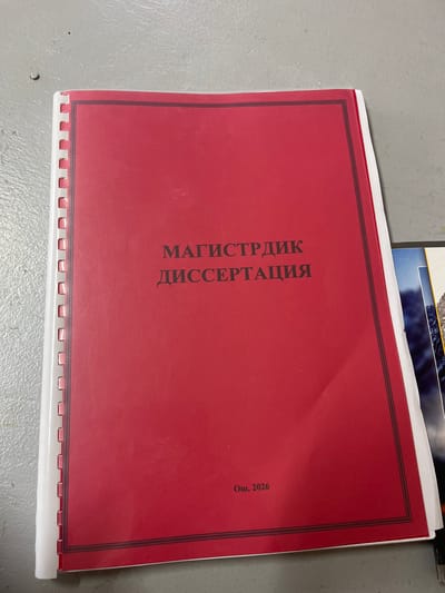 A red binder with spiral binding on the left side lies on a gray concrete surface. The binder has a black border and contains Russian text that reads "МАГИСТРДИК ДИССЕРТАЦИЯ" in the center and "Ош, 2026" at the bottom. To the right of the red binder, another binder with a picture of mountains is partially visible. The image is taken from a slightly overhead angle. There are no people in the image. The environment appears to be indoors, possibly a storage area or office space. The binder suggests the document is related to a master's thesis in the city of Osh, Kyrgyzstan, in the year 2026.