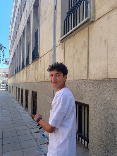 An outdoor, eye-level, medium shot captures a young man with curly dark hair, wearing a white t-shirt and a dark wristwatch, as he looks over his left shoulder towards the camera. He is holding a clear plastic bottle in his right hand, with his thumb pointing upwards. The man stands on a paved sidewalk next to a long, multi-story building with a light-colored stone facade. The building features rows of rectangular windows with black metal bars. To the left of the man, the sidewalk extends away from the camera, revealing a few parked cars and a bicycle in the distance. The sky is clear and blue. The scene appears to be in Vidin, Bulgaria, given the architectural style and context.