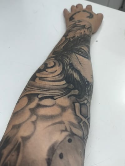The image displays a close-up, angled view of a person's forearm and hand, extensively covered in black and grey tattoos. The tattoos are intricate, featuring swirling patterns, what appears to be a face or figure with flowing hair, and geometric shapes. There are also circular elements, some appearing as dots or voids within the design. The skin has some visible hair, indicating it's a human arm. The tattoos are in various stages of shading, with some areas appearing dark and solid, while others are more faded or blended. The hand, visible at the top of the frame, also bears a tattoo resembling a laurel wreath or similar circular design. The subject is resting their arm on a white surface, likely a table, which is the primary element of the background. The lighting is neutral, suggesting an indoor setting. There is no visible text in the image. The overall impression is of a detailed, possibly artistic, tattoo sleeve. The context provided indicates the location as Kolpino, Russia, but there are no specific visual cues in the image to confirm this.