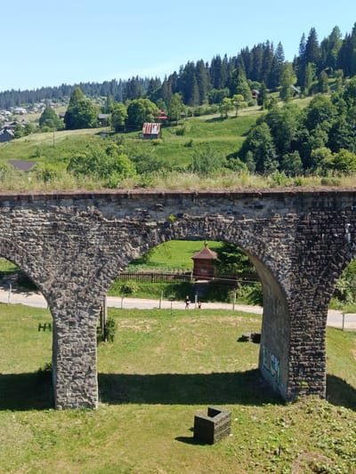 This is a photograph taken in the city of Vorokhta, Ukraine, capturing a section of a large stone viaduct that spans across a grassy area. The viaduct, constructed of rough-hewn grey stones, features prominent arches. In the foreground, the viaduct's pillars cast long shadows on the grass. A small, dark, square stone object sits on the grass near the right pillar.

Through one of the viaduct's arches, a small, red, wooden structure resembling a chapel or a historical monument is visible. A wooden fence runs behind it. A paved road is visible beyond the fence, and two individuals, appearing to be children or women, are walking along the grassy area beside the road, casting shadows.

The background depicts a hilly landscape covered in lush green grass and dense forests of coniferous trees. Several houses and buildings, with visible roofs, are scattered across the hills, indicating a residential area. The sky is clear and blue, suggesting a sunny day. The overall scene portrays a rural or semi-rural mountainous setting with a significant historical architectural element.