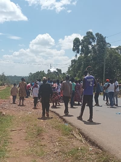 A group of approximately 20-30 individuals is observed walking along and beside a paved road in Chevaywa ward, Kenya, on a clear, sunny day. The road, which features a distinct dark asphalt surface, has a dirt and grassy shoulder on its left side. Several plastic bottles are visible as litter on the roadside.
The individuals are predominantly adults, with some children present. They are casually dressed; one male in the foreground wears a purple sleeveless shirt with the number "2" on the back, and another wears a dark long-sleeved shirt and trousers. Several women are wearing patterned dresses. The group appears to be generally moving in the same direction, with some walking on the paved surface and others on the dirt shoulder. One individual in the mid-ground holds a white flag attached to a pole.
The background features numerous green trees, some notably tall, suggesting a vegetated or semi-rural environment. Overhead, power lines are visible against a bright blue sky with scattered white cumulus clouds. Distant hills or elevated terrain are discernible on the horizon.