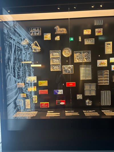 This is a photograph of a museum display in Kilchberg (ZH), Switzerland. The display features a black background with various artifacts and memorabilia related to the Nestlé company. The items are arranged in rows and columns, with some items cast in silver or pewter, and other original packaging. The bottom of the display shows an image of a Nestlé factory. The artifacts include vintage chocolate molds, chocolate bar packaging, and promotional materials. The numbers 1, 2, 3, and 4 are visible, indicating the location of particular items on the display. Labels are visible for brands and products such as Cailler, Lindt, and Nestlé.
