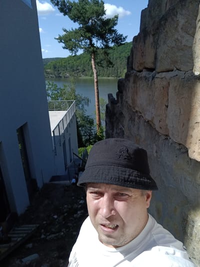 A man wearing a black bucket hat and a white t-shirt is taking a selfie outdoors. He is positioned to the right of the frame, with his face filling the foreground. Behind him, to his left, is a modern building with a light gray facade and a white railing on an upper level. The building appears to be under construction or renovation, with visible dirt and debris on the ground. To the right of the man is a rough stone wall. In the background, a body of water, likely a lake, is visible, surrounded by lush green trees and hills under a bright blue sky with scattered clouds. The scene is bathed in sunlight, casting shadows. The approximate location is identified as Doksy, Czechia.