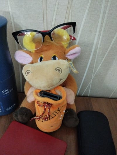A stuffed toy cow wearing oversized black-rimmed glasses sits on a wooden surface. The cow has yellow horns and pink ears. It is holding an orange pouch with red and gold string. A dark blue bottle with the text "ESTEL" and "9% ОКСИГЕНТ" is to the left of the cow. A red rectangular object is in the foreground to the left, and a black rectangular object is to the right. The background is a textured beige wall. The scene is indoors and appears to be on a table or desk. The location is not specified in the image or the provided crops.