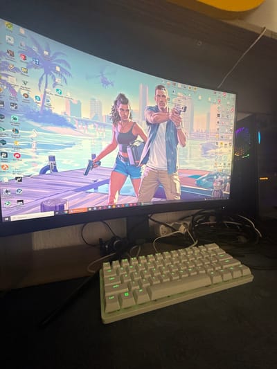 A monitor displays an image reminiscent of a Grand Theft Auto loading screen, depicting a man and a woman holding guns against a backdrop of a city skyline and palm trees, likely inspired by Miami. The monitor screen is also filled with icon shortcuts including recognizable logos for games and software. A white mechanical keyboard sits to the right of the monitor, and various cables are visible, suggesting a gaming setup. Above the monitor, a partially visible computer case with a Gigabyte logo is seen. The surface underneath is dark, and a hint of a yellow object is visible on the left. The location of Breza, Bulgaria is not verifiable with the available information.