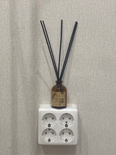 A brown glass diffuser bottle with black reeds stands on a white electrical outlet adapter. The bottle has a label that reads "PARFUME HOME" and some unreadable text below it. The outlet adapter is mounted on a textured, light-colored wall. The background is a plain, unadorned wall with a subtle woven pattern. There are no people or animals present. The scene appears to be an indoor domestic setting. The location in Dolgoprudny, Russia, is not determinable from the image itself.