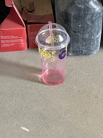 The image depicts a close-up of a pink plastic cup with a clear plastic dome lid and a straw. The cup has a yellow circular design with a spokes pattern and a purple "C" shape printed on it. To the left of the cup are parts of red and brown cardboard boxes. The box on the left has white text printed on it. To the right of the cup is a large, dark, textured plastic container. The scene is set on a grey, textured surface that appears to be pavement or concrete. The background is blurred, but suggests an outdoor or industrial setting. The image does not contain any discernible information about Strumivka, Ukraine.