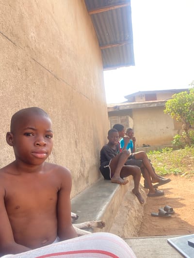 The image captures an outdoor scene in Idiroko, Nigeria, under bright daylight. A young, bald Black boy is seated in the foreground, facing the camera with a neutral expression. He is shirtless and wears white and red striped lower garments. Behind him, a white dog's head and foreleg are visible, resting on the ground. To the right, three additional young Black boys are seated on a low, concrete bench. The boy closest on the bench wears a black t-shirt and dark shorts with a white stripe, looking towards the camera with a slight smile; he is barefoot. The other two boys are partially obscured, one in a light blue shirt looking away, and another in dark clothing. Two pairs of dark slide sandals are visible on the reddish-brown dirt ground near the bench. The background consists of a textured, light brown building wall with a corrugated metal roof overhang. Beyond this, additional buildings with corrugated metal roofs and light walls are visible, alongside green foliage from a bush or tree. The sky is bright and appears clear. A dark object, possibly a laptop or tablet, is partially visible in the bottom right corner.