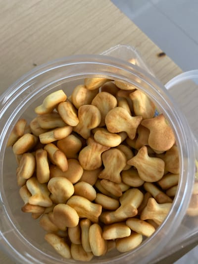 A close-up, high-angle shot shows a clear plastic container filled with goldfish-shaped crackers. The crackers are golden brown and piled high within the container, which is placed on a light-colored wooden surface. The background is slightly blurred, showing more of the wooden surface and a hint of white to the right, possibly another container lid. The lighting is natural, coming from above and slightly to the left, casting subtle shadows on the crackers and the container's rim. There is no indication of a specific location in Ta', Indonesia.