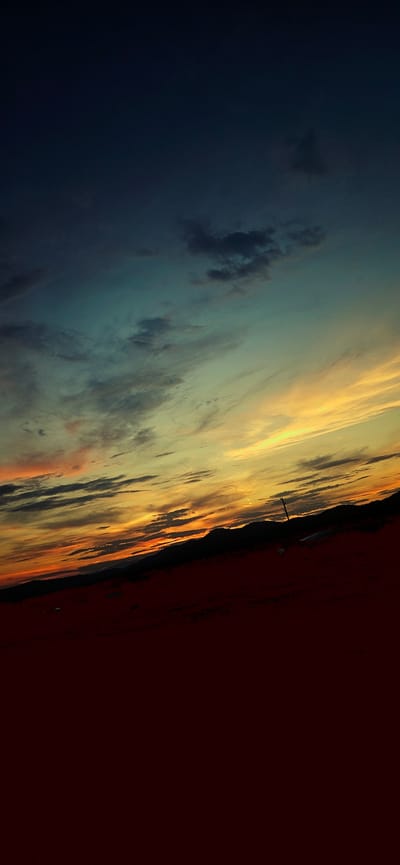This is a vertical, slightly tilted photograph of a sky with a dramatic sunset over a dark landscape. The upper portion of the image is a deep, dark blue transitioning into teal and then a bright yellow and orange band where the sun is setting. Wispy clouds are visible throughout the sky, catching the light in various shades. Below the sky is a dark, silhouetted mountain range. In the foreground, the ground is a deep red or brown, also appearing silhouetted and indistinct. A thin, vertical dark object, possibly a stick or pole, is present in the middle ground, silhouetted against the sky. The image appears to be taken from an elevated position looking down towards the horizon. The location is identified as Momin Prohod, Bulgaria, suggesting a natural landscape setting, likely mountainous or hilly. The lighting indicates dusk or twilight.