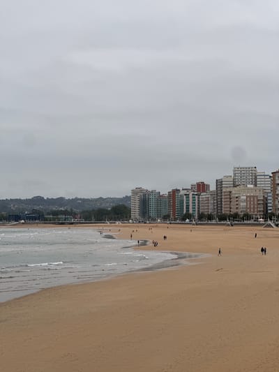 The image shows an aerial view of San Lorenzo Beach in Gijón/Xixón, Spain, on a cloudy day. The beach is a wide expanse of golden sand bordering the Bay of Biscay. Waves are visible along the shoreline. A row of high-rise buildings lines the edge of the beach, indicating a densely populated urban area. Some individuals are scattered along the beach, likely walking or engaging in leisure activities. A green landscape is situated behind the buildings. The sky is overcast with gray clouds, suggesting the weather is cool.