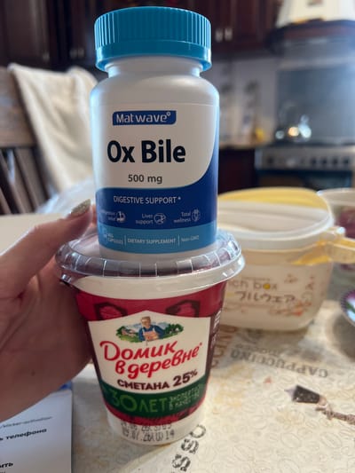 A close-up, vertically oriented, slightly blurry shot shows two food products and a hand. On the left is a white plastic bottle with a blue cap, labeled "Matwave Ox Bile 500 mg." Below the main label are smaller text lines indicating "DIGESTIVE SUPPORT," "Indigestion relief," "Liver support," and "Total wellness," along with icons. To the right of the bottle is a red and white plastic container with a clear lid, displaying Russian text that reads "Домик в деревне СМЕТАНА 25%" (Domik v derevne Sour Cream 25%). The lid has a printed design of a farmer. The background appears to be a table with a patterned tablecloth, possibly with coffee bean motifs and text like "CAPPUCCINO." There's also a glimpse of a kitchen appliance in the upper left corner. A hand with glittery nail polish is holding the container. The scene seems to be indoors, possibly a kitchen or dining area in Krasnoe-na-Volge, Russia. The overall impression is casual, with the products laid out on a surface.