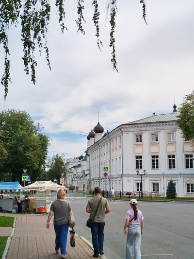 This image depicts a street scene in Yaroslavl, Russia. Three people are walking away from the camera down a brick-paved sidewalk. The person in the foreground is a woman with blonde hair wearing a gray t-shirt and blue pants, carrying a beige bag over her shoulder and a patterned bag in her right hand. Behind her walks a man in a green t-shirt and blue jeans, with a brown bag slung across his chest. A young girl in a pink t-shirt and light blue jeans, wearing a white cap and an orange bag across her body, walks in the street to the right of the man.

The scene appears to be set in a historic part of the city, with a grand white building with numerous windows and decorative architectural details lining the right side of the street. Several dark-domed church towers rise above the building. Trees are visible on the left side of the street, and in the foreground, there are market stalls with tents and umbrellas. One tent has text that appears to say "МОРОЖЕНОЕ" (ice cream). Another umbrella is white with a circular pattern.

The sky is partly cloudy, suggesting it might be daytime, perhaps late morning or afternoon. The weather appears mild, as people are dressed in casual, light clothing. The overall mood of the image is relaxed and everyday. A pedestrian crossing sign is visible on the left side of the street.