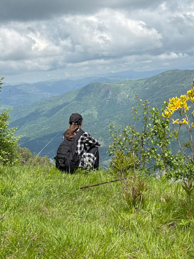 A person wearing a black baseball cap and a black and white checkered shirt and pants, carrying a black backpack, sits on a grassy hillside overlooking a vast, mountainous landscape. The person is facing away from the camera, looking towards the distant mountains. Yellow flowers bloom on a bush in the foreground to the right of the person. The sky is cloudy, with dramatic grey and white clouds. The location is the mountainous region near Nizhnyaya Teberda, Russia.