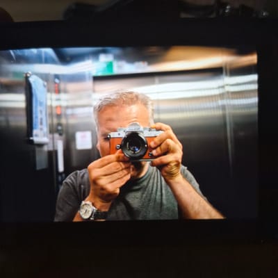 The image shows a man taking a selfie in an elevator. He is holding a vintage-style camera with an orange leatherette finish and a silver top plate. The man has short, graying hair and is wearing a gray t-shirt and a silver wristwatch. The elevator interior appears to be made of brushed stainless steel. On the left wall of the elevator, there is a dispenser with a blue and white label, and a red illuminated button. The overall impression is that of a modern, clean elevator car. It is not possible to determine the specific location within Başakşehir, Turkey based on the provided image.
