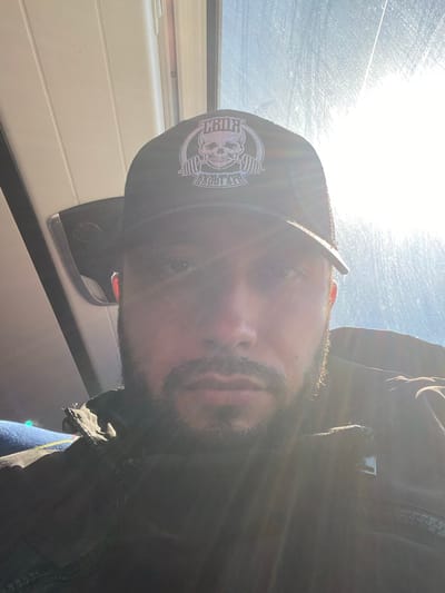 A man wearing a black baseball cap with a skull logo is pictured in a selfie taken indoors. The cap is dark with a light-colored logo featuring a skull and barbell. The man has a beard and is looking towards the camera. The background shows interior elements of what appears to be a vehicle, with sunlight streaming in from a window, creating lens flare and obscuring details of the environment. The lighting suggests it is daytime. There is no visible indication of the location being in Koskolovo, Russia.