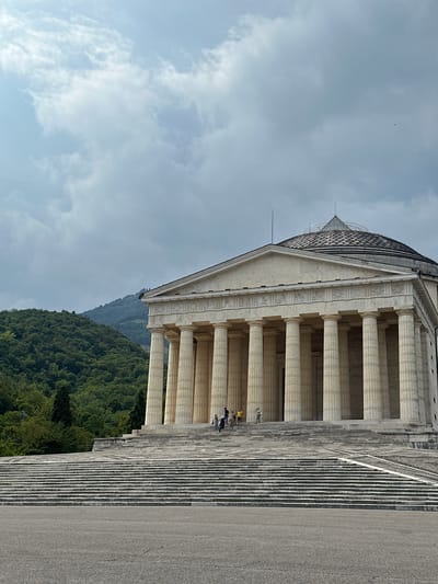 The image shows a large, Neoclassical-style building with a portico supported by numerous columns. The building is set against a backdrop of lush green hills under a cloudy sky. A wide, long staircase leads up to the entrance of the building. Several people are visible on the stairs and at the entrance, appearing to be tourists. The architecture suggests a historical monument or a public building. The location is identified as the Temple of Antonio Canova in Possagno, near Crespano del Grappa, in the province of Treviso, Italy. The building's design, with its prominent dome and classical facade, is characteristic of the works of Antonio Canova, an influential Italian sculptor. The surrounding landscape indicates a rural or semi-rural setting, likely in the pre-alpine region of Veneto.