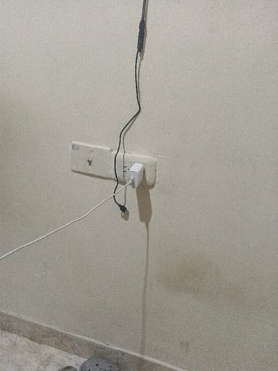 A close-up interior shot displays a beige wall with a white electrical outlet, into which a white charger is plugged. Two black cords descend from above the outlet, while a white cord extends from the charger towards the left. The wall shows slight imperfections and discoloration. Near the bottom of the frame, a section of tiled floor and a portion of what appears to be a gray shoe are visible.