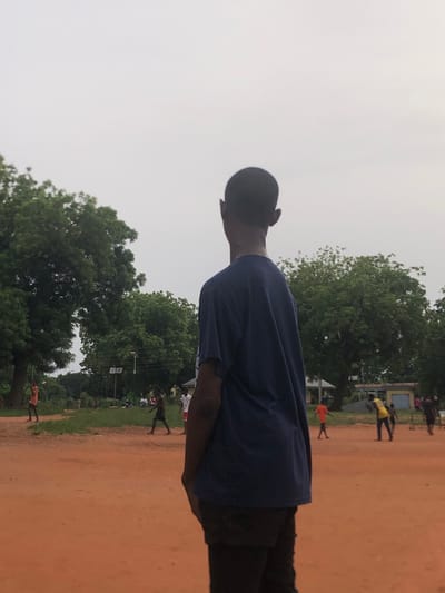 This media file captures an outdoor scene, likely in Nsugbe, Nigeria, under an overcast sky.

The main subject, positioned in the foreground, is a young person, presumably male, with short dark hair. They are seen from behind, wearing a navy blue short-sleeved t-shirt and dark trousers, looking towards the right side of the frame.

The setting is a large, open field covered in reddish-brown dirt or sand. The background features a line of dense, green trees, suggesting a natural or semi-rural environment. The sky is a uniform light grey, indicating an overcast day without direct sunlight.

In the mid-ground and background, several other individuals are visible. One person stands out in a bright yellow shirt and dark pants, near a light-colored building or structure. Further back, a smaller figure in an orange top is present. Other people are scattered across the field and near the treeline, appearing to be walking, standing, or engaging in casual activities. Utility poles or power lines are faintly visible among the trees.

The image depicts a casual outdoor gathering or daily life in an open community space, possibly a schoolyard or a local field. The time of day cannot be precisely determined but appears to be either early morning or late afternoon due to the diffuse light, or simply a cloudy day. There is no visible text in the image.