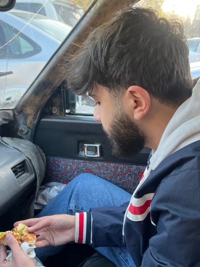 Within the Kafr Sousa Municipality, Syria, a male individual is seated inside a vehicle, observed from the chest up in profile, looking downwards. He has dark, unkempt hair and a dark beard, and is wearing a white hooded sweatshirt beneath a navy blue jacket with white and red striped cuffs and accents, along with blue denim jeans. His hands hold a partially consumed slice of pizza, wrapped in white paper, featuring visible green pepper, black olive, and cheese toppings.
The immediate environment is the interior of a vehicle, displaying signs of wear and age. The left A-pillar is visible, appearing worn and possibly damaged, next to a cracked windshield through which other vehicles and daylight are discernible. A dashboard or side panel section to the left includes an air vent. The vehicle's door panel to the right features a patterned fabric insert with red and light blue elements and a chrome door handle. The external environment, seen through the vehicle's windows, shows other parked or stationary vehicles, suggesting an outdoor urban setting.