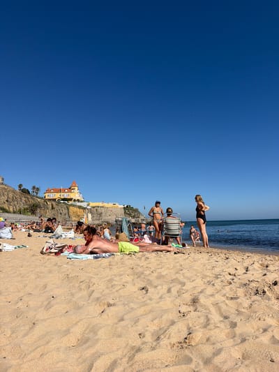 The image depicts a sun-drenched beach scene in Cascais, Portugal. The foreground is dominated by textured sand with footprints, indicating recent activity. A clear, deep blue sky stretches across the upper portion of the frame. In the mid-ground, a group of people are scattered across the sand, some lounging on towels and beach chairs, while others are in the water. The ocean is a calm blue, meeting the sandy shore.
Further back, buildings with distinctive architecture, characteristic of coastal Portugal, are visible on a cliffside overlooking the beach. These buildings are colorful, with terracotta roofs and yellow facades, suggesting a resort or town area. Some palm trees are interspersed among the buildings and on the cliffs.
The people are dressed in swimwear, engaged in typical beach activities like sunbathing and swimming. Their poses and the overall atmosphere suggest a leisurely summer day. The sun is high in the sky, indicating it is likely midday or early afternoon. The weather appears to be warm and clear, with no clouds visible. The mood is relaxed and vacation-like. There is no visible text in the image.