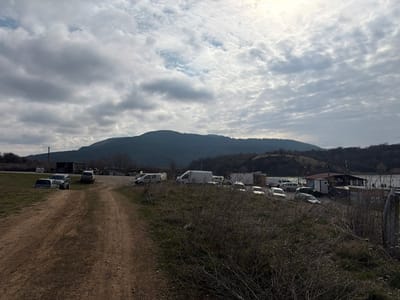 This image depicts a rural or semi-rural scene in Cherna Gora, Bulgaria, characterized by an overcast sky with scattered altocumulus clouds. The foreground features a dirt track with visible ruts extending from the bottom center, flanked by dry, sparse grass and brush. In the midground, to the left of the track, are several parked vehicles, including at least one sedan partially embedded in the ground. To the right of the track, a larger informal parking area contains a cluster of numerous vehicles, predominantly cars and white vans, alongside multiple small, utilitarian structures, one with a brown roof, positioned near the edge of a large body of water. The background is dominated by a dark, tree-covered mountain range on the horizon, with rolling hills closer to the midground, some featuring sparse deciduous trees. No individuals are visible.