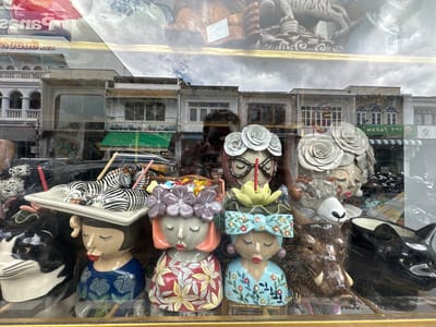 This media file captures a display of decorative ceramic figures and other items through a glass window, likely a shopfront in Phuket City Municipality, Thailand. The subjects include several human-like ceramic figurines with closed eyes and distinctive facial features, some adorned with floral headpieces and bows. Other objects visible are ceramic animals, including a black and white cat, a wild boar, a ram, and two zebras resting on a platter. The background reflections suggest an outdoor scene with buildings and a cloudy sky.

The display appears to be static, with no discernible activity or event taking place beyond the presentation of merchandise. Notable details include the intricate floral patterns on the figurines' clothing, the textured detailing of the animal figures, and the reflections of the external environment on the glass. The lighting suggests it might be daytime, with overcast conditions due to the cloudy sky.

There is some visible text in the image, primarily appearing as reversed lettering on what looks like signage reflected in the glass. One prominent reflection shows what appears to be "ice shop" or similar lettering in white on a green background. Other reflected text is less legible due to the glare and distortion from the glass. The overall impression is one of a quirky and artistic display of decorative items for sale.