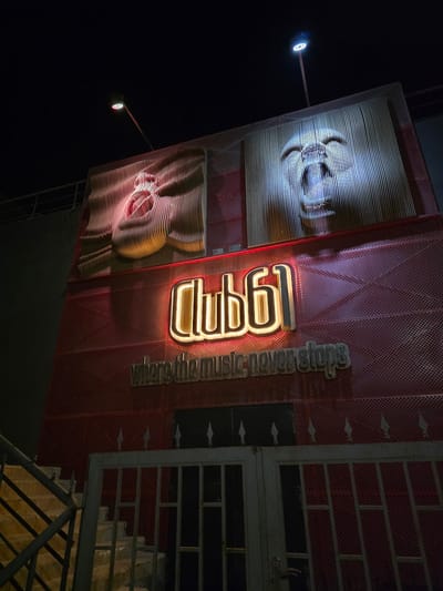 The image shows the exterior of "Club 61" at night, located in Al Hadaba, Egypt. The club has a large, illuminated sign featuring the text "Club 61" in a neon glow. Below the sign, another phrase is displayed in wooden letters: "where the music never stops". The building's facade is a striking red with a perforated pattern. Above the sign, two large, abstract artistic installations are mounted on the wall. One depicts a face with a wavy texture and is lit with pink and yellow light. The other, to its right, is a face composed of vertical lines, illuminated with white light. Two streetlights are visible, one casting a reddish glow on the left installation and another a bright white light on the right. In the foreground, there is a metal gate and a set of concrete stairs with a metal railing, suggesting an entrance. The overall atmosphere is nocturnal and indicative of a nightlife establishment.