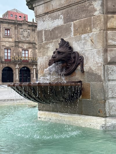 A lion-head fountain spouts water into a pool. The fountain is attached to a stone wall. Behind the fountain, a building with the name "CONMIMU" on it can be seen. This is located in the city of Gijón/Xixón, Spain.