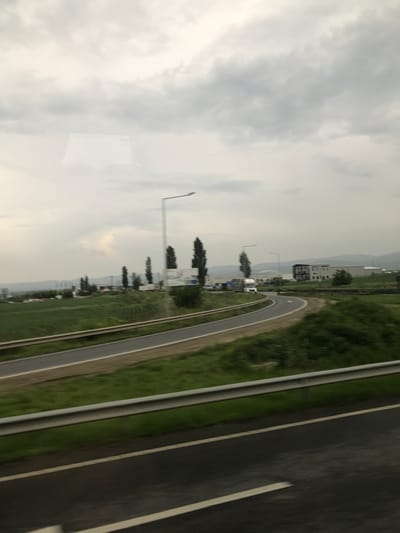 The image depicts an overcast daytime scene of a highway and an adjacent industrial/commercial area near Nicolae Bălcescu, Romania. In the foreground, a dark asphalt road with distinct white lane markings extends from the bottom of the frame, bordered by a horizontal, two-bar metal guardrail positioned across the lower third. Beyond this guardrail, another section of asphalt road curves towards the right, flanked by vibrant green grassy verges.

In the midground, a curving road leads towards a cluster of low-rise buildings. A white semi-truck is visible on this curved road, heading towards the buildings. Several tall, slender trees with dark foliage are interspersed within the green landscape. Two modern streetlights with long, cantilevered arms rise vertically, providing illumination. A large, rectangular white sign with dark text, possibly reading "DESCOVA" in part, is situated among the trees on the right side. Various other vehicles are sparsely parked or moving near the buildings in the mid-distance.

The background features additional commercial or industrial structures, primarily white or light-colored, with one multi-story building displaying dark windows. Distant rolling hills or mountains are visible under a uniformly grey, cloudy sky that dominates the upper half of the image. A faint, rectangular reflection of an internal object is discernible in the upper-left quadrant, suggesting the photograph was captured through a window. No individuals are clearly identifiable in the scene.