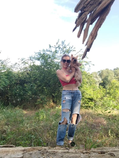 A woman is holding a small dog in her arms in an outdoor setting. She is wearing a pink strapless top and ripped blue jeans. The dog is light brown and appears to be a Yorkshire Terrier. They are standing in front of a dense green bush with trees in the background. The sky is partly cloudy. A piece of weathered wood hangs from the top right of the frame. The ground is covered with dry grass and a fallen log is in the foreground. The image does not contain any contextual information that would allow for the determination of the location being Fyodorovskaya, Russia.